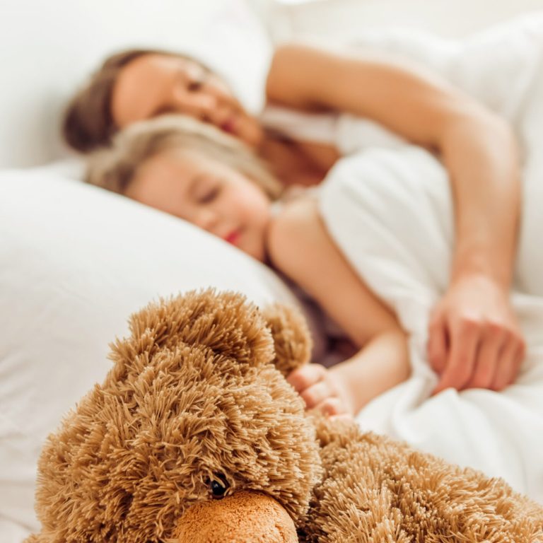 Sweet little girl and her beautiful young mother are sleeping with a teddy bear in bed at home